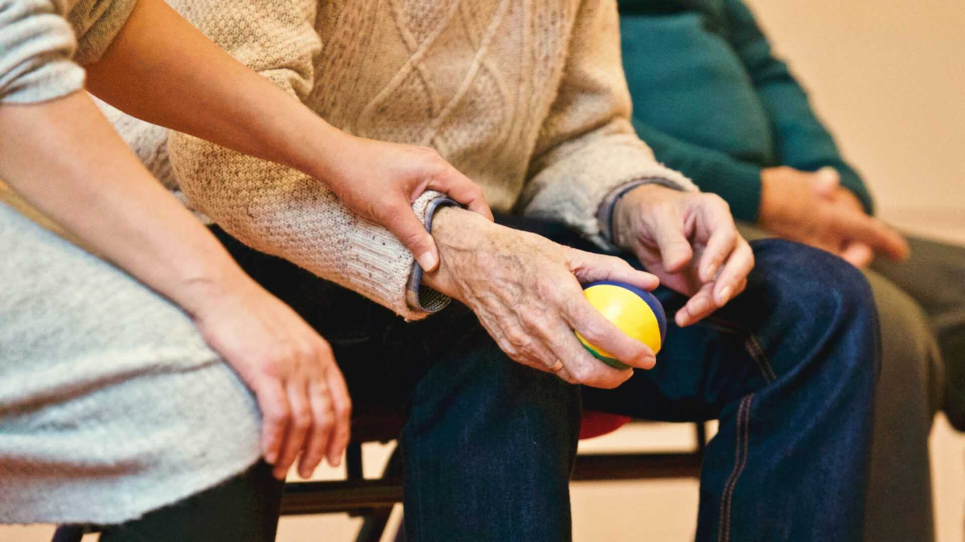 Ein alter Mann hält einen Ball, eine Pflegekraft unterstützt ihn dabei.