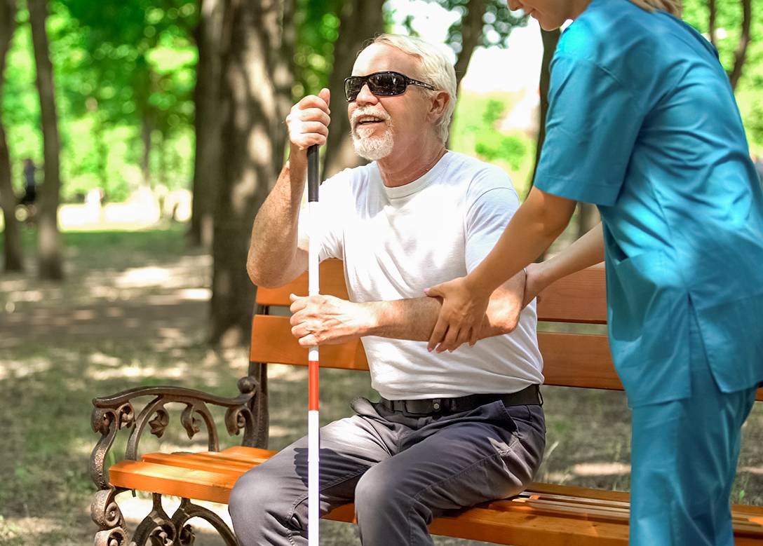 Ein Mann mit Blindenstock sitzt auf einer Parkbank. Eine junge Pflegerin hilft ihm.
