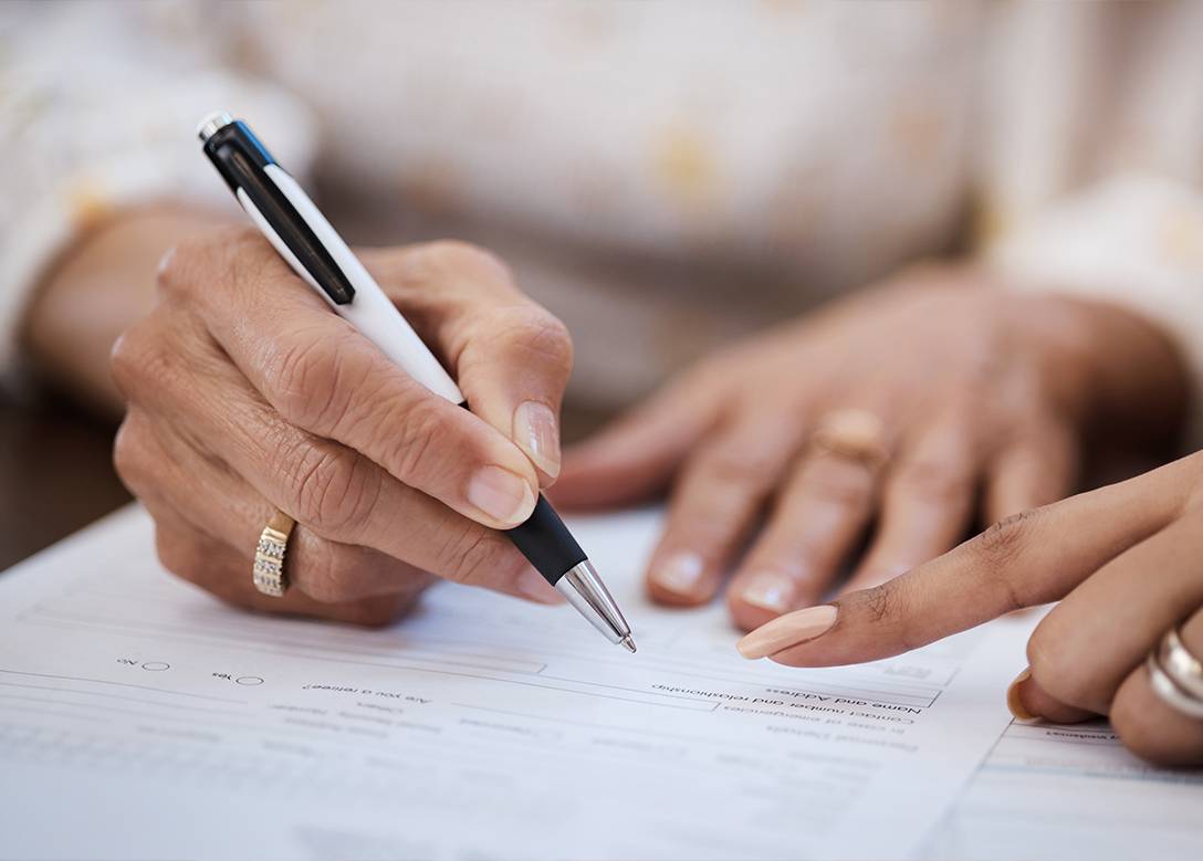 Eine Person unterzeichnet ein Dokument. Die Hand einer Frau weist auf eine Passage im Text hin.