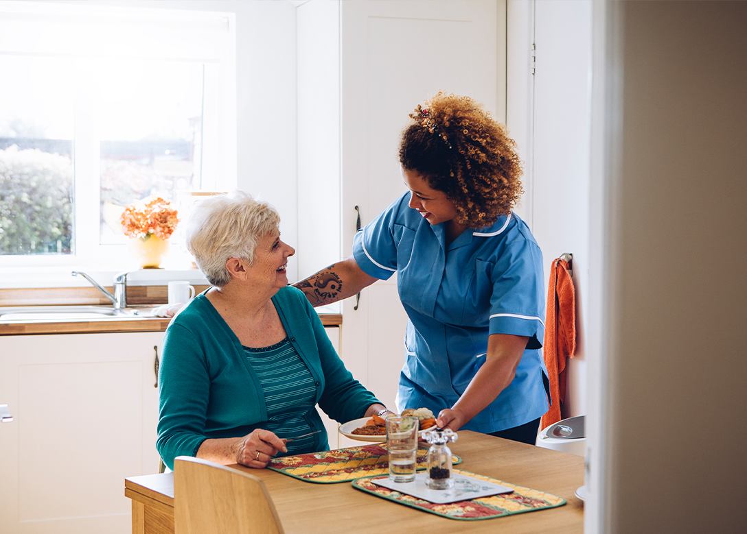 Eine weibliche Pflegekraft tellt einer älteren Frau einen Teller mit Essen auf den Tisch.