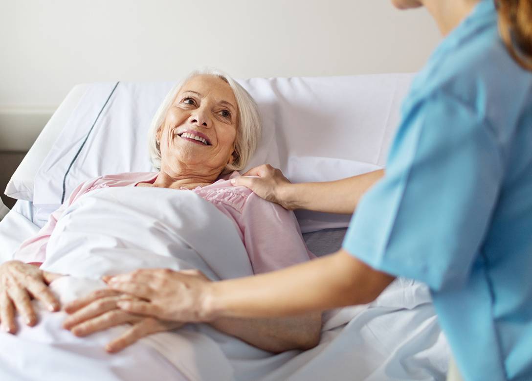 Eine ältere Frau liegt im Bett, lächelt ihre Pflegekraft an.