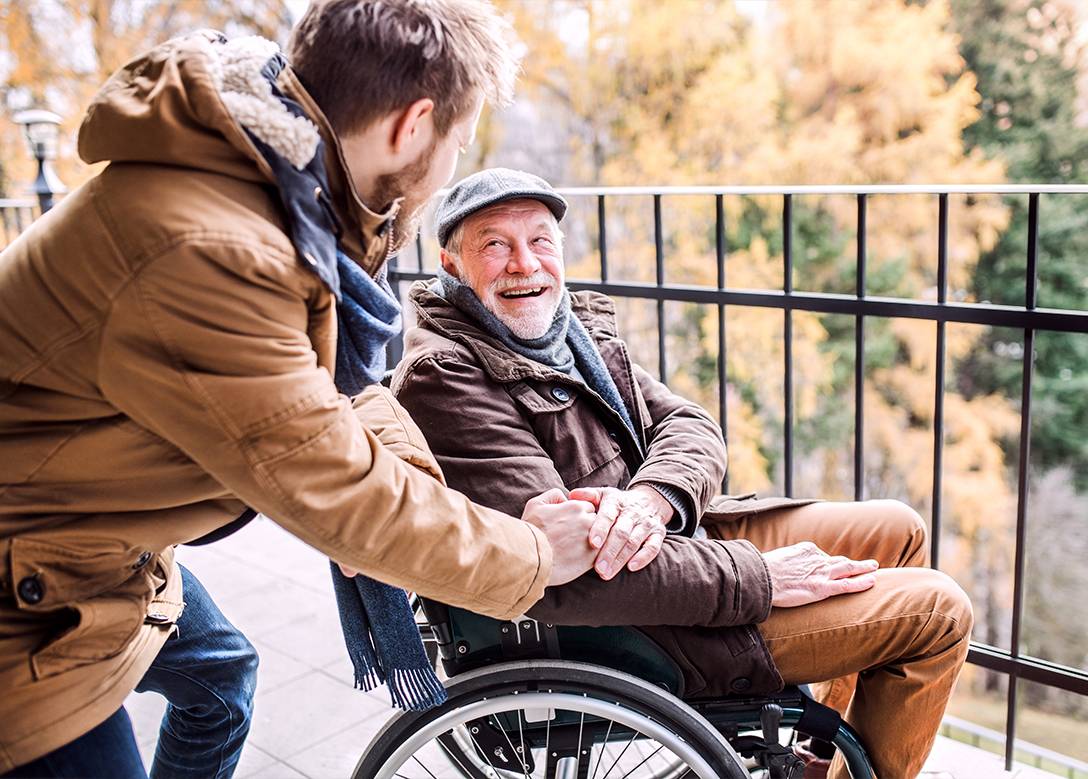 Ein älterer Mann in einem Rollstuhl unterhält sich mit einem jüngeren Mann. Beide lachen.